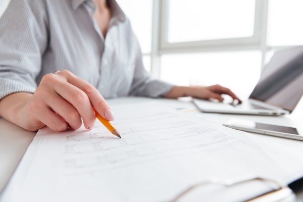 Close up portrait of a woman holding pencil over paper documents while sitting and working with laptop computer at home